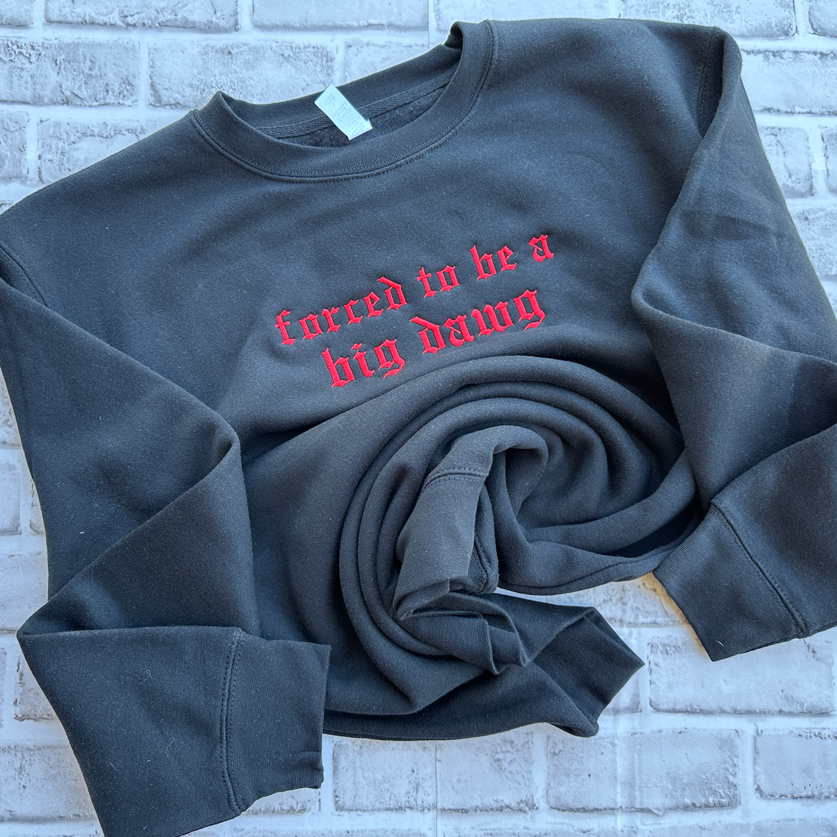 Black crewneck sweatshirt featuring red Old English embroidered text across the chest reading “forced to be a big dawg,” shown flat on a white background.
