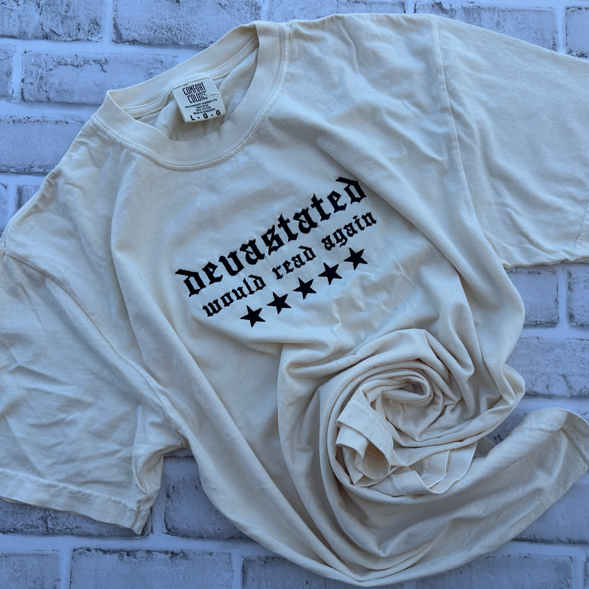 Comfort Colors T-shirt featuring black Old English embroidered text reading “devastated” with “would read again” underneath and five black stars across the chest.