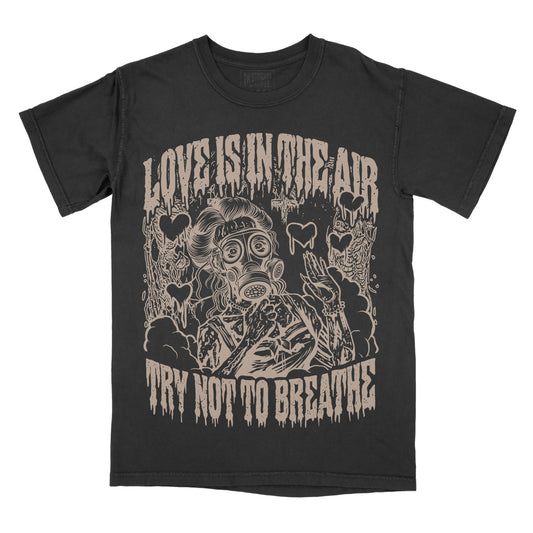 A black short-sleeve graphic t-shirt featuring a distressed beige print of a person in a gas mask surrounded by skulls and hearts, with the text 'LOVE IS IN THE AIR' above and 'TRY NOT TO BREATHE' below.