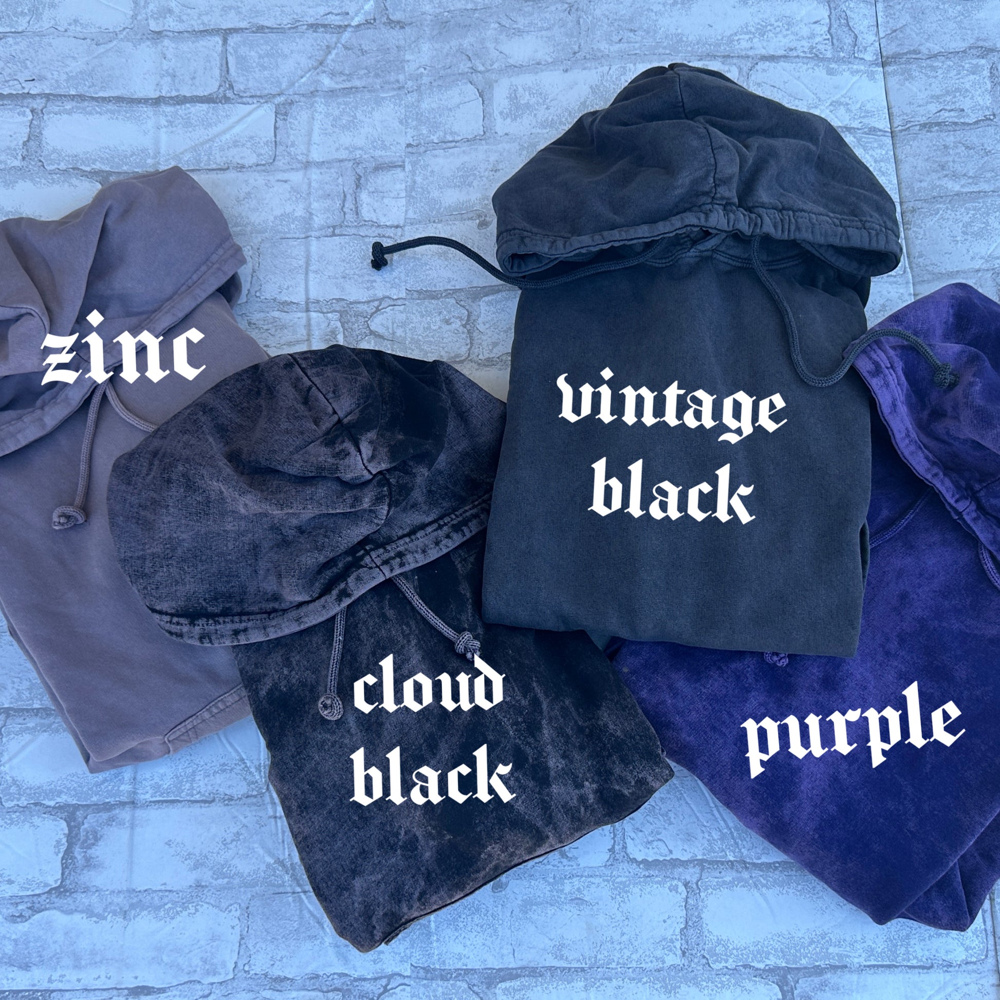 Four folded mineral washed hooded sweatshirts shown on a light brick surface, labeled zinc gray, cloud black, vintage black, and purple to display available color options.