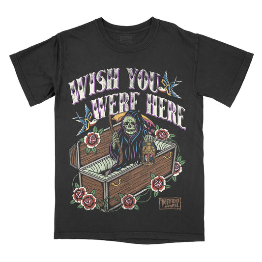 Black garment-dyed graphic t-shirt featuring the phrase "Wish You Were Here" with an original Grim Reaper illustration emerging from a coffin, surrounded by roses, swallows, and traditional tattoo-inspired artwork