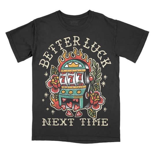 A black short-sleeve t-shirt with a vintage-style tattoo-style graphic of a slot machine, roses, and the text 'BETTER LUCK NEXT TIME' on the front, shown against a plain white background.