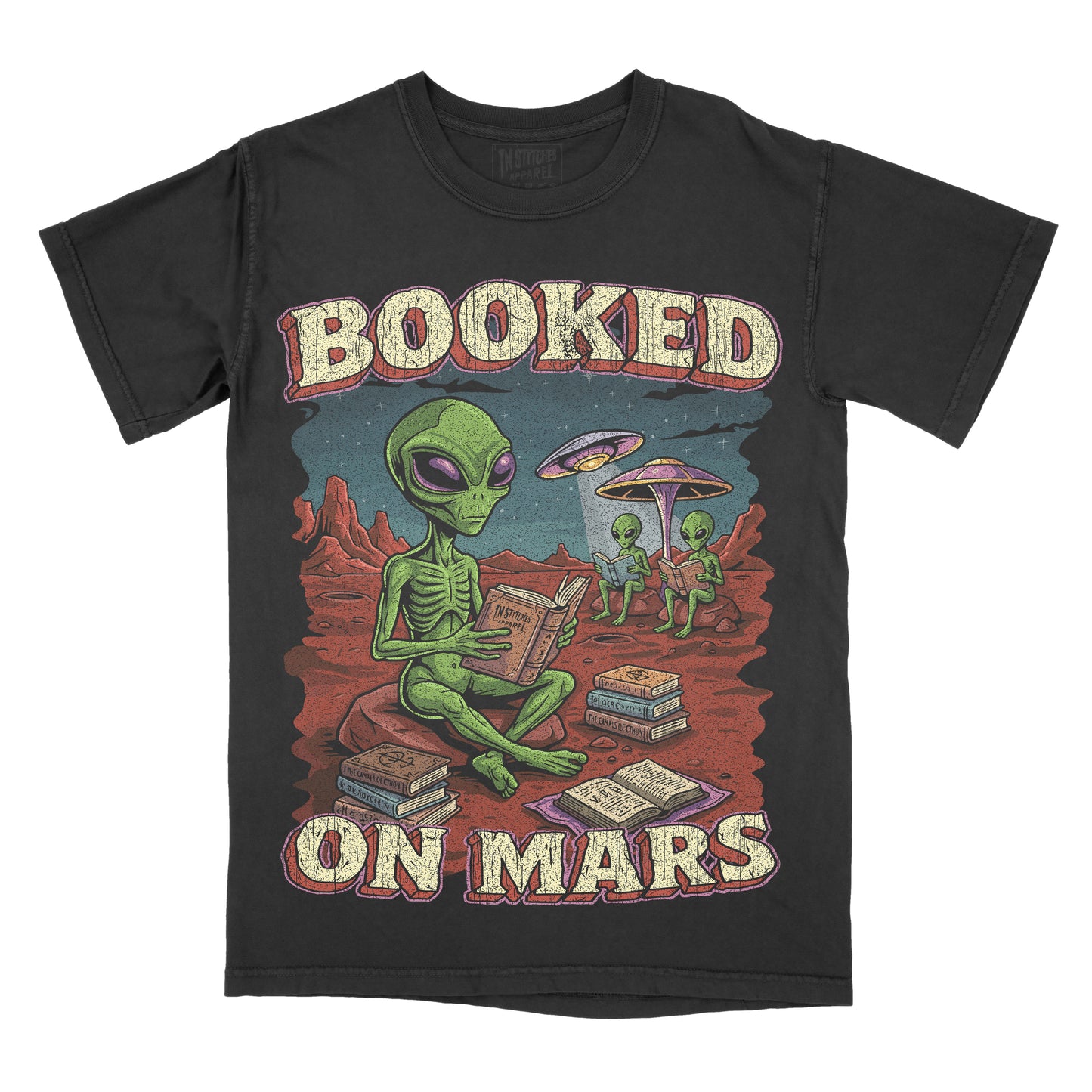 A black short-sleeve graphic t-shirt featuring a retro-style illustration of green aliens on a red desert planet reading books, with the text 'BOOKED ON MARS' in a distressed cream font.