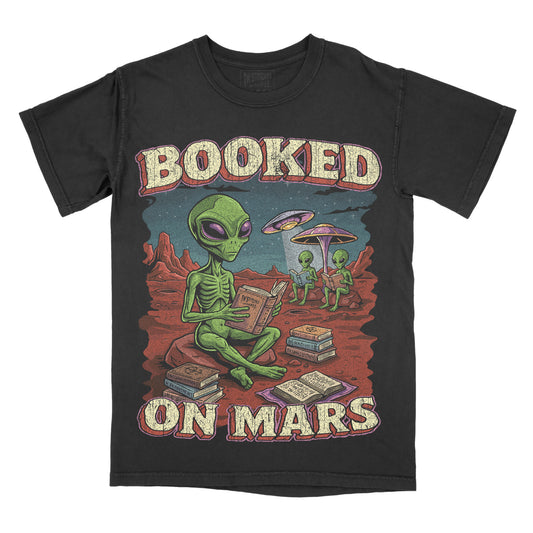 A black short-sleeve graphic t-shirt featuring a retro-style illustration of green aliens on a red desert planet reading books, with the text 'BOOKED ON MARS' in a distressed cream font.