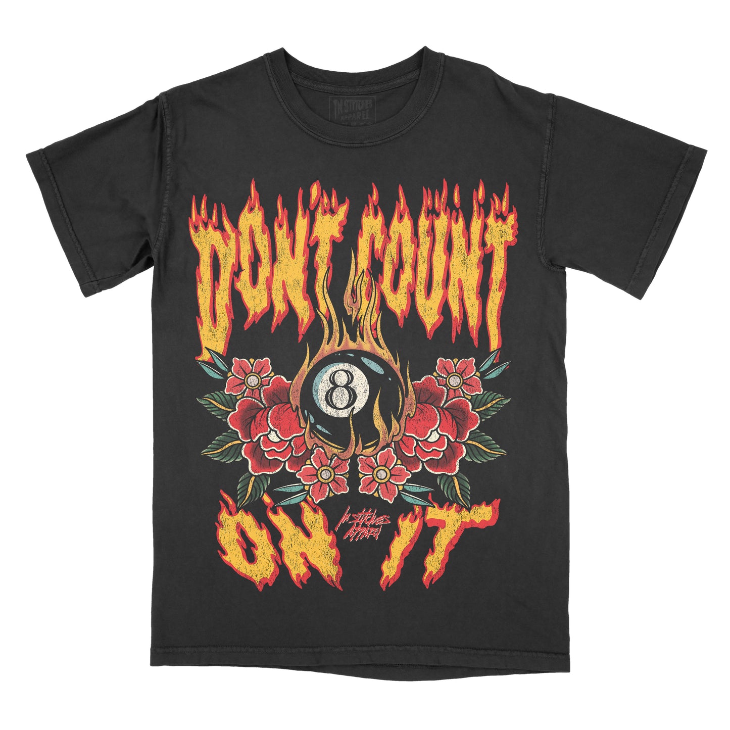“Black vintage-style t-shirt featuring flame-styled lettering that reads ‘Don’t Count on It.’ The design shows a black 8-ball surrounded by red flowers and green leaves, with flames rising around the graphic. The artwork uses red, yellow, green, and tan tones in a bold, tattoo-inspired style. ‘In Stitches Apparel’ branding appears below the design.