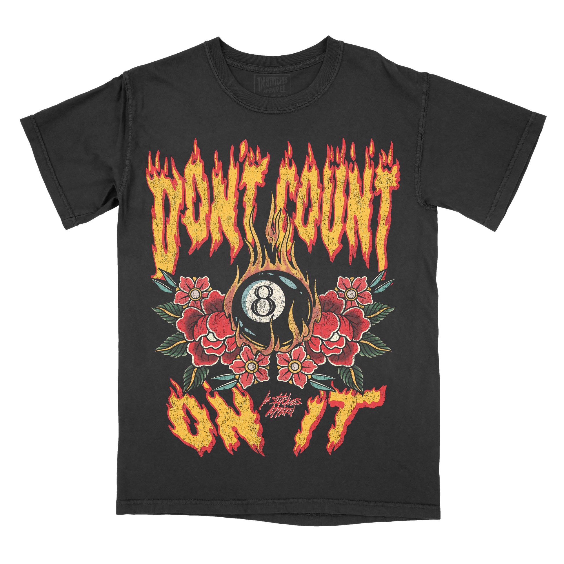“Black vintage-style t-shirt featuring flame-styled lettering that reads ‘Don’t Count on It.’ The design shows a black 8-ball surrounded by red flowers and green leaves, with flames rising around the graphic. The artwork uses red, yellow, green, and tan tones in a bold, tattoo-inspired style. ‘In Stitches Apparel’ branding appears below the design.