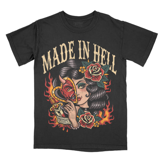 Black Comfort Colors graphic T-shirt featuring a vintage tattoo-style illustration of a woman holding a devil heart mask with roses, flames, and the phrase "Made in Hell."