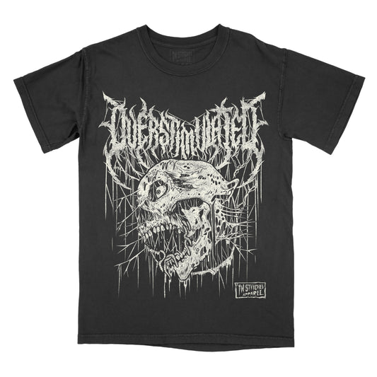 A black short-sleeve graphic t-shirt featuring a white distressed metal logo that reads 'overstimulated' above a detailed white skull illustration with dripping paint effects. A small 'In Stitches Apparel' brand label is visible on the lower right of the graphic.