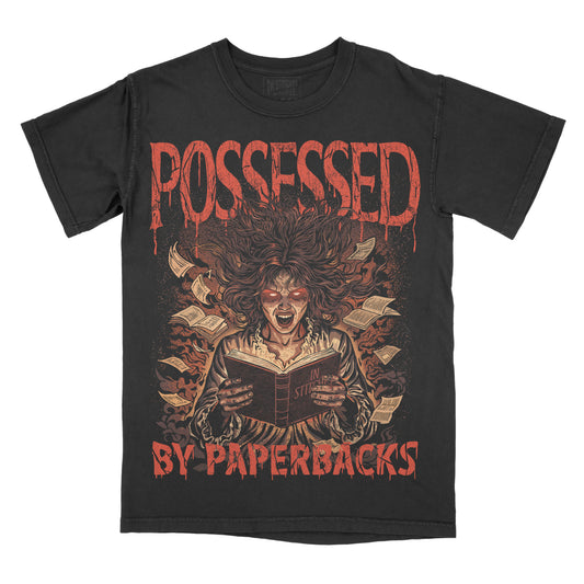 A black short-sleeve graphic t-shirt featuring a horror illustration of a screaming woman reading a book surrounded by books, with the text 'POSSESSED BY PAPERBACKS' in a dripping red font.