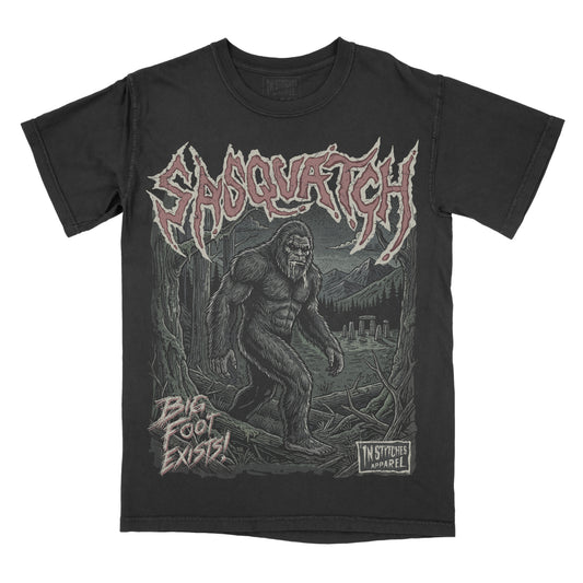 A black short-sleeve graphic t-shirt featuring the Sabotage's 'Big Foot Exists!' artwork. The design shows a detailed illustration of Bigfoot in a forest with the word sasquatch in a stylized font at the top and a 'In Stitches Apparel' logo at the bottom right.