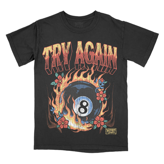 A black short-sleeve graphic t-shirt featuring a distressed design with the words 'TRY AGAIN' in orange-red flame lettering above an 8-ball billiard graphic surrounded by flames and red flowers. A small 'In Stitches Apparel' brand label is visible near the bottom right of the graphic.